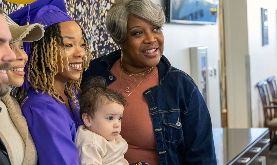Four adults and a baby pose and smile for a photo at a graduation celebration. One woman wears a purple cap and gown, while others stand close together in a festive indoor setting.