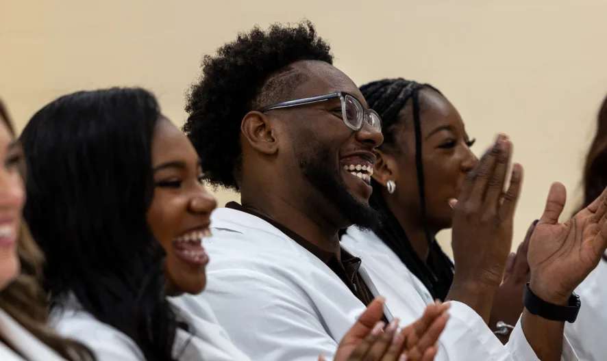 Five people in white coats sit in a row, smiling and clapping. The group appears to be celebrating or attending a formal event, such as a ceremony. The background is plain and light-colored.