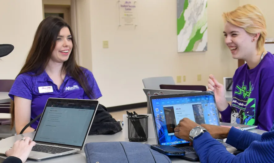 Four students sit around a table with laptops, talking and smiling. Two students wear purple shirts, one wears a black hat, and one wears a blue sweater. They appear to be collaborating in a bright, casual setting.