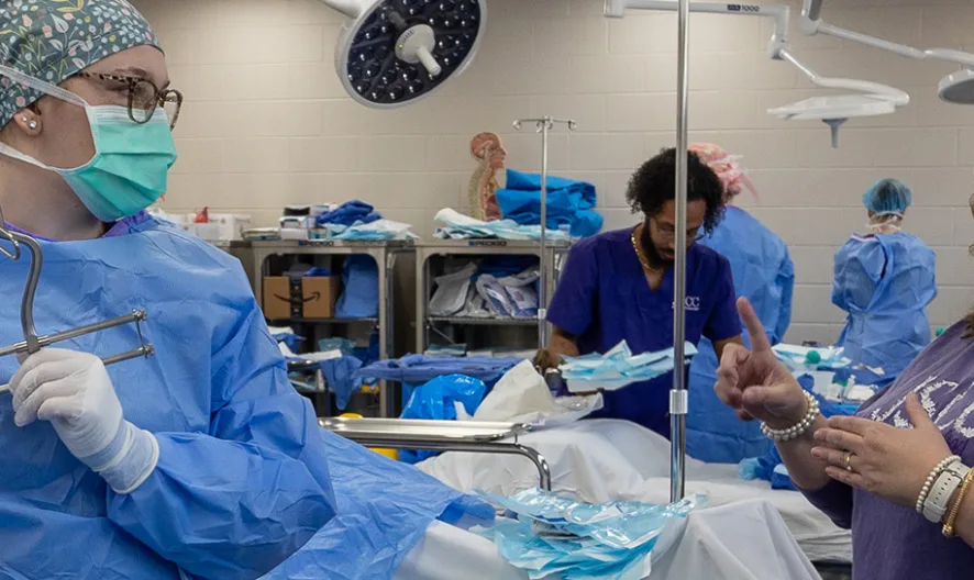 A woman in scrubs and a mask holds surgical instruments while talking to another woman in a purple shirt inside a medical training room, with people and medical supplies in the background.