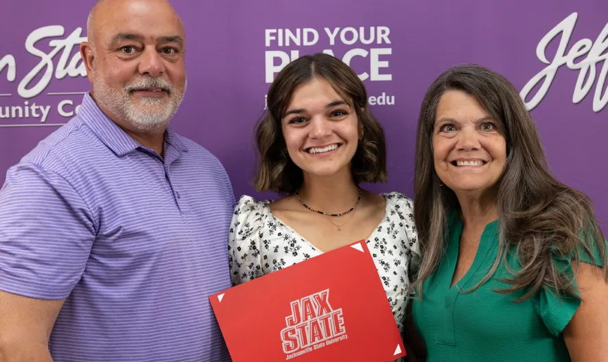 Three people stand smiling in front of a purple Jefferson State Community College banner. The person in the center holds a red Jax State folder.