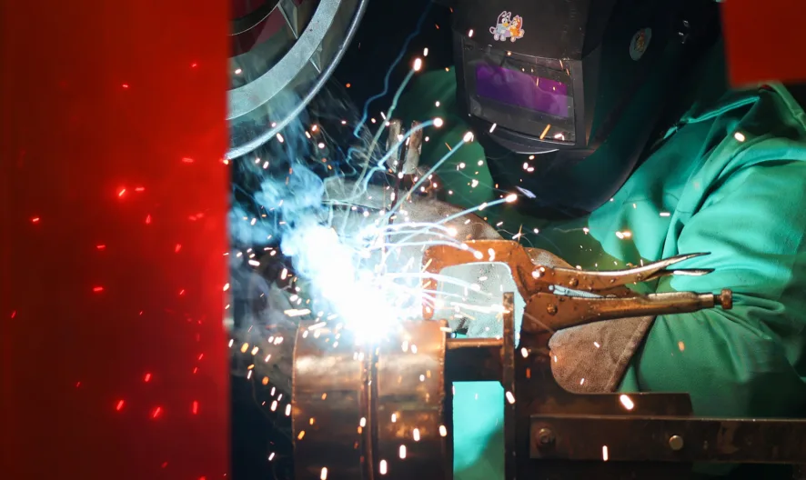 A person wearing protective gear and a welding helmet uses a welding torch, creating bright sparks and blue light in a workshop setting.