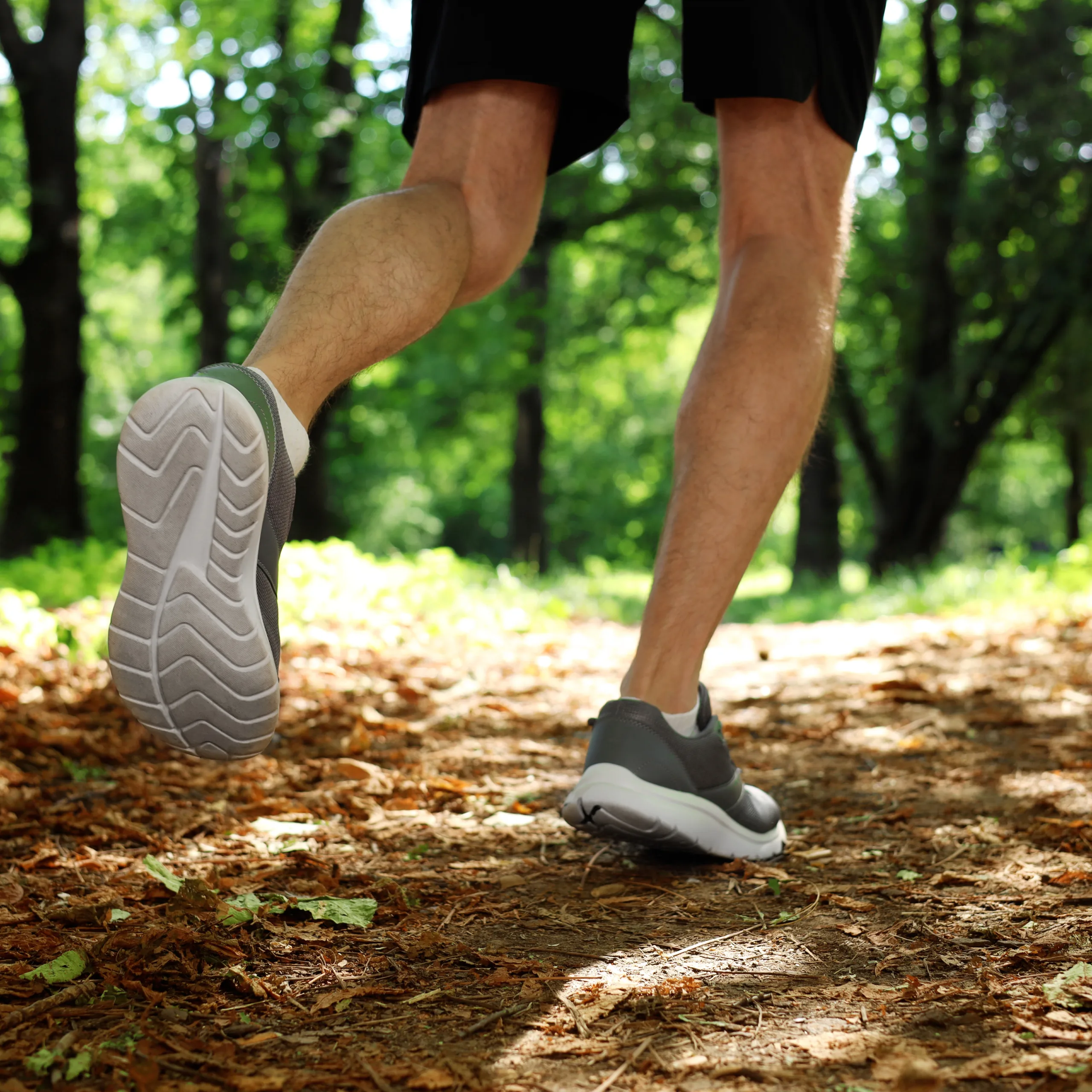 A person wearing shorts and gray running shoes jogs on a forest trail covered in leaves and wood chips, surrounded by green trees and sunlight filtering through the canopy.