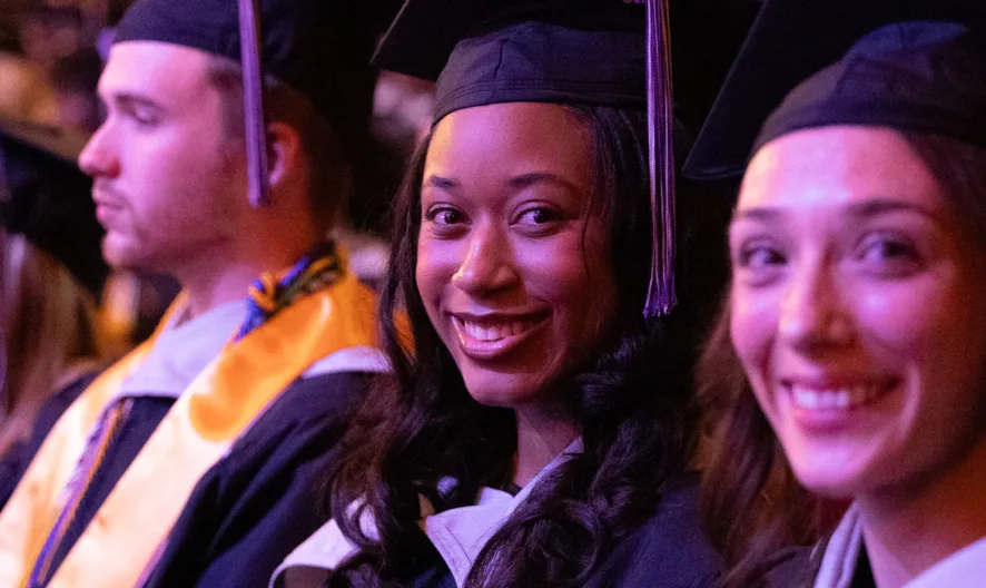 A group of students in graduation caps and gowns sit in rows at a ceremony. Two women in the foreground are smiling at the camera, while other graduates sit beside them, facing forward.