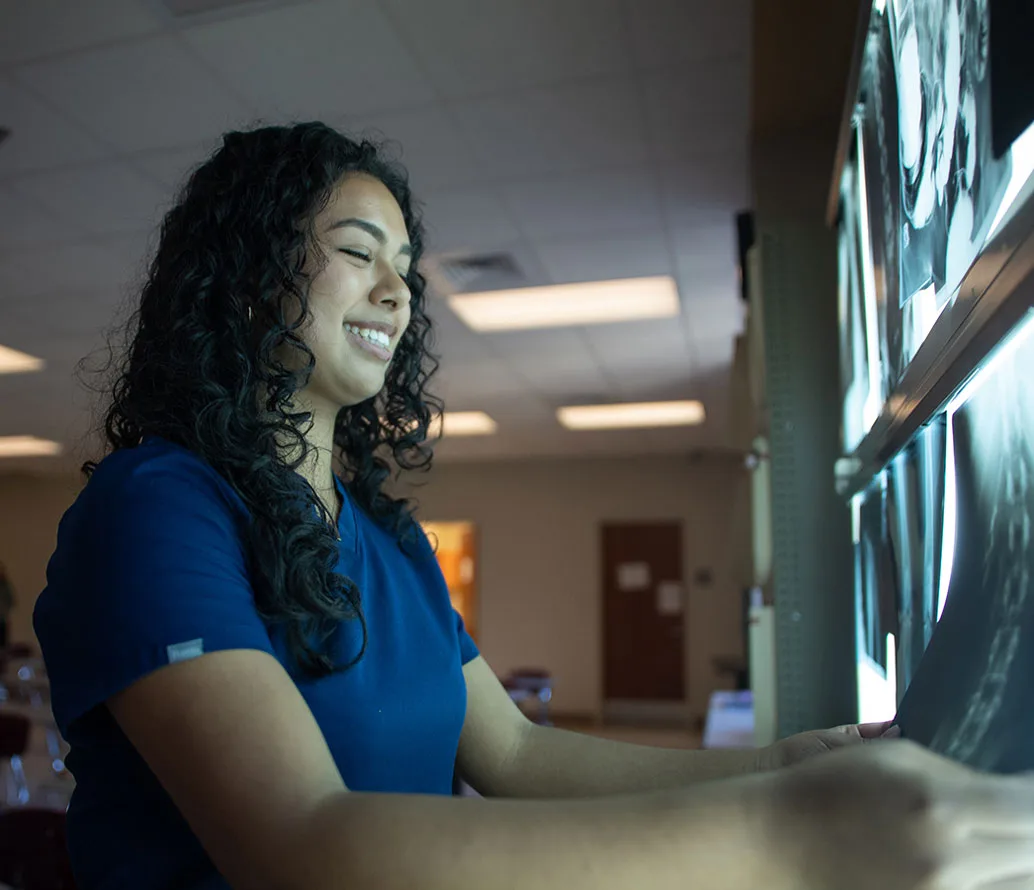 A woman in blue scrubs smiles while examining X-ray images on a lighted display board in a medical setting.