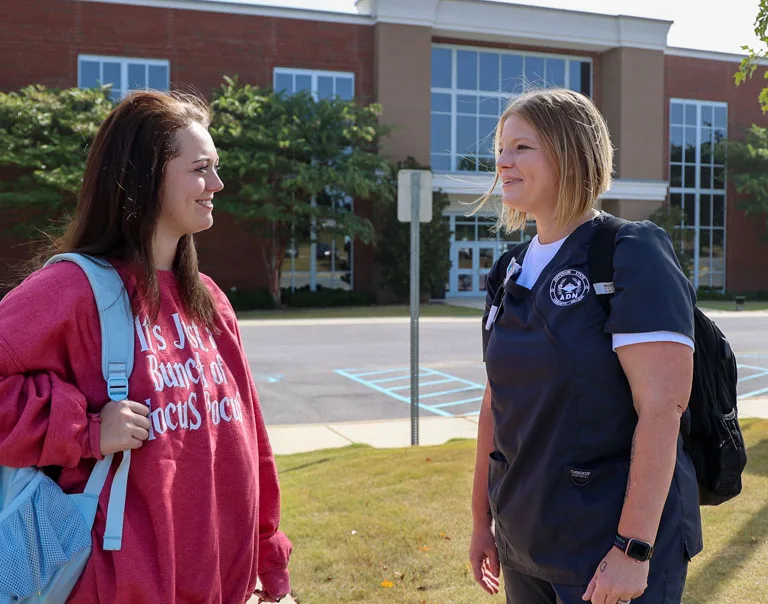 Two women stand outside a building, smiling and talking. One wears a red sweatshirt and carries a light blue backpack; the other wears navy scrubs and a black backpack. The building has large windows and trees along the front.