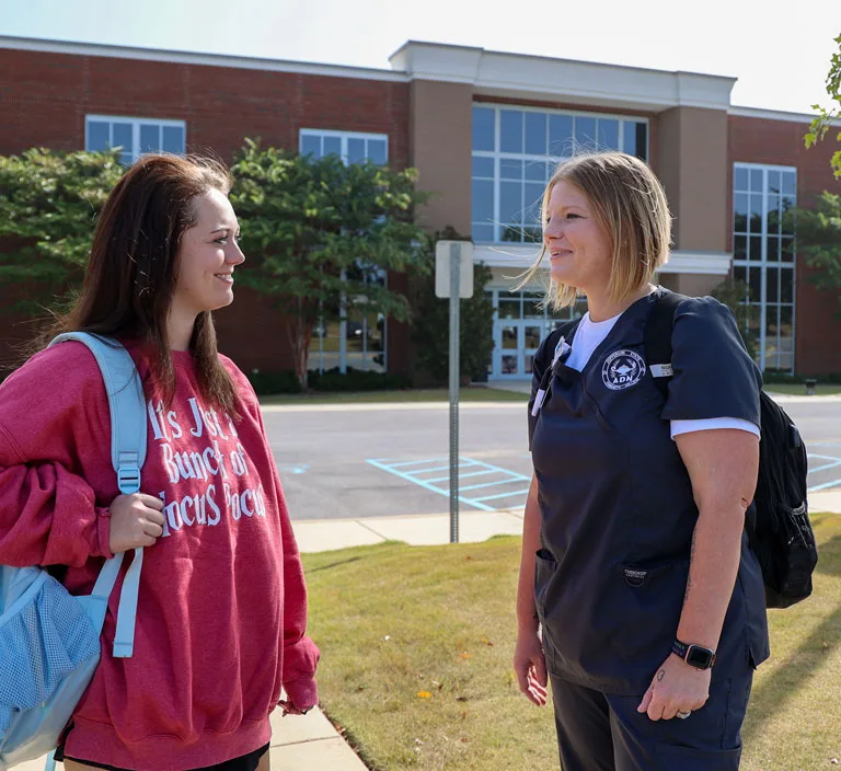 Two women stand outside a building, smiling and talking. One wears a red sweatshirt and carries a light blue backpack; the other wears navy scrubs and a black backpack. The building has large windows and trees along the front.