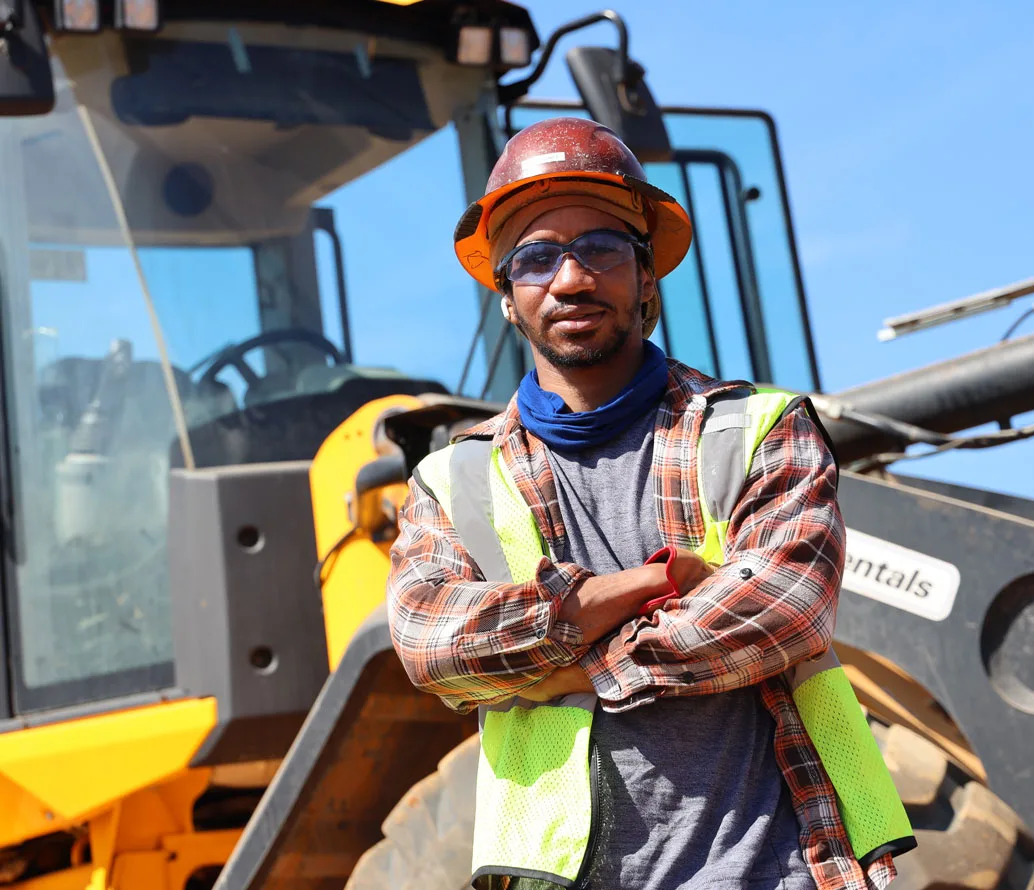 A construction worker wearing a hard hat, safety glasses, and a reflective vest stands with arms crossed in front of heavy machinery on a sunny day.