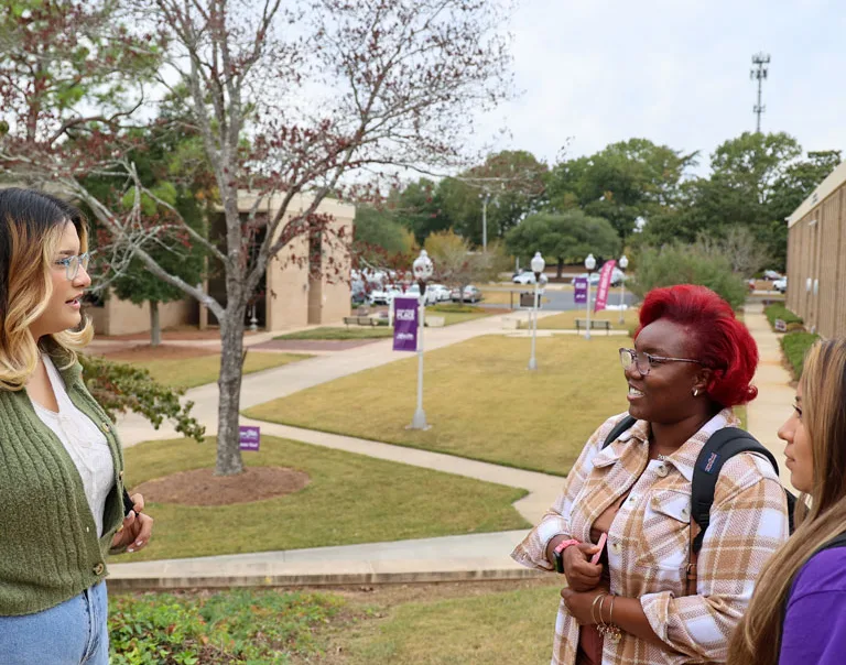 Three students stand outdoors on a college campus, engaged in conversation. Two students face the camera, one with red hair and glasses, and the other with long brown hair. A third student stands opposite them.