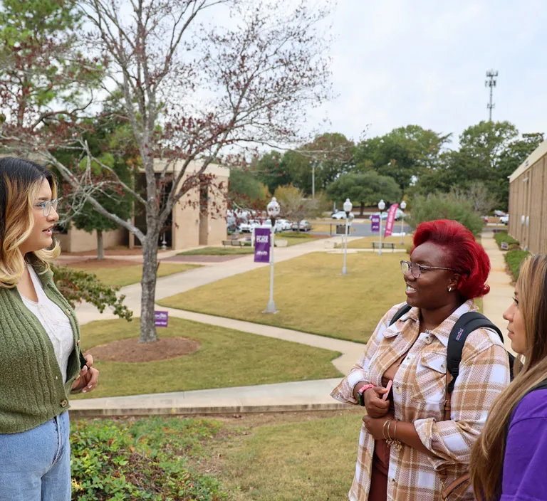Three students stand outdoors on a college campus, engaged in conversation. Two students face the camera, one with red hair and glasses, and the other with long brown hair. A third student stands opposite them.