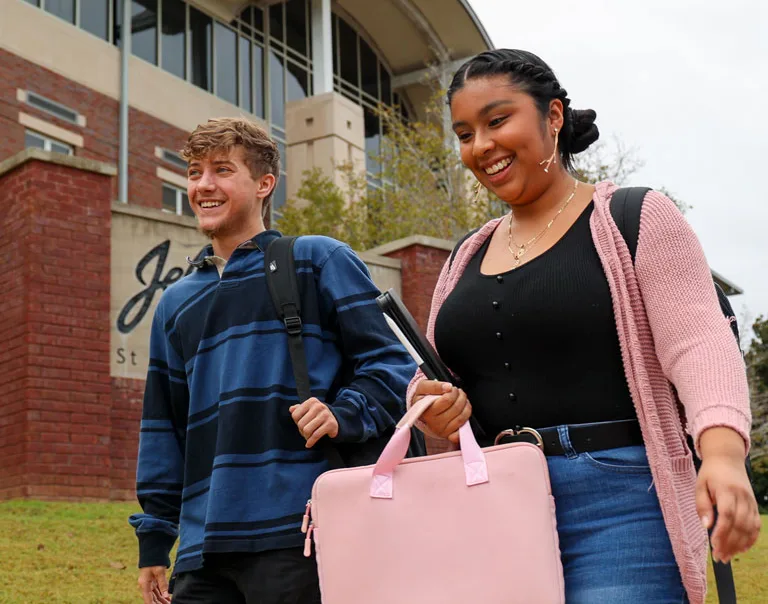 Two college students, a smiling young man and woman, walk outside a campus building. The woman carries a pink bag and a notebook, while the man carries a backpack. The building and grass lawn are visible in the background.