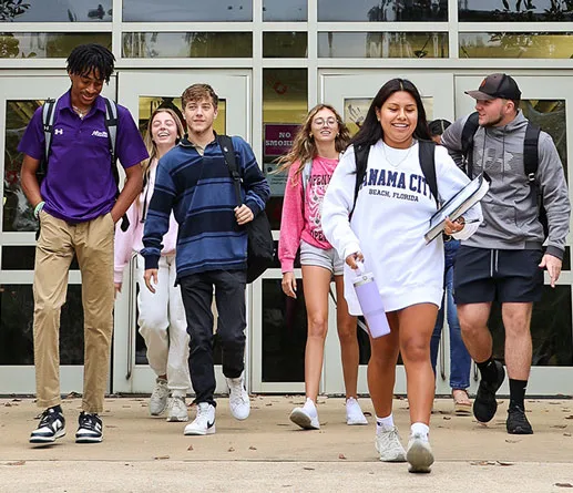 A group of six smiling students walk outside a school building carrying backpacks and books. They appear happy and relaxed, dressed in casual clothing. The background shows glass doors and windows.