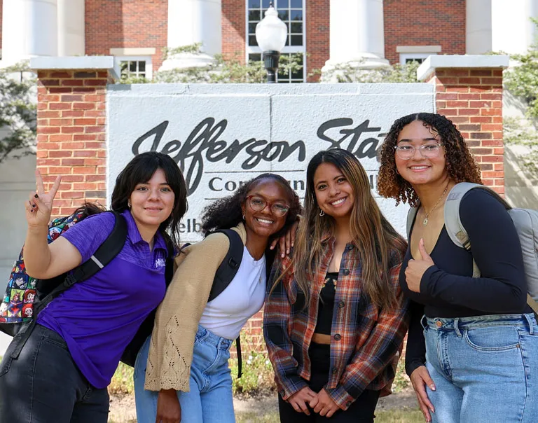 Four smiling students with backpacks stand in front of a Jefferson State Community College sign, posing for a group photo on a sunny day.