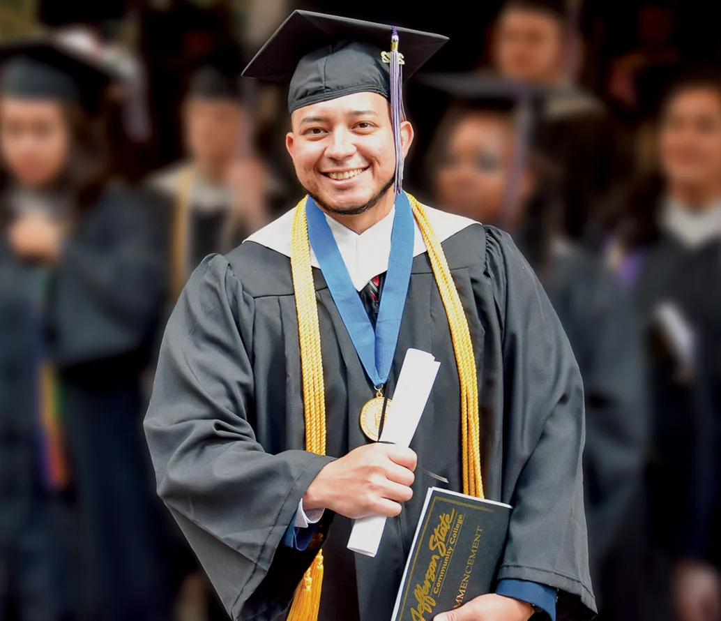 A smiling graduate in a cap and gown holds a diploma and certificate, wearing honor cords and a medal, with other graduates blurred in the background.