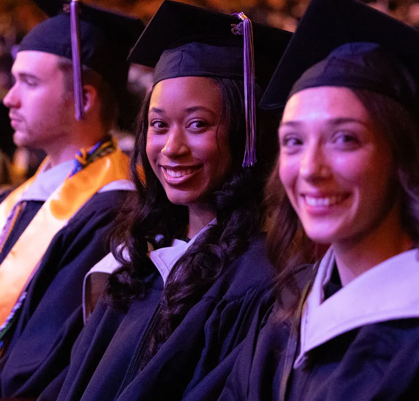 Three graduates in caps and gowns sit in a row at a ceremony. The young woman in the middle smiles at the camera, while the other two graduates, a man and a woman, sit on either side of her and look forward.