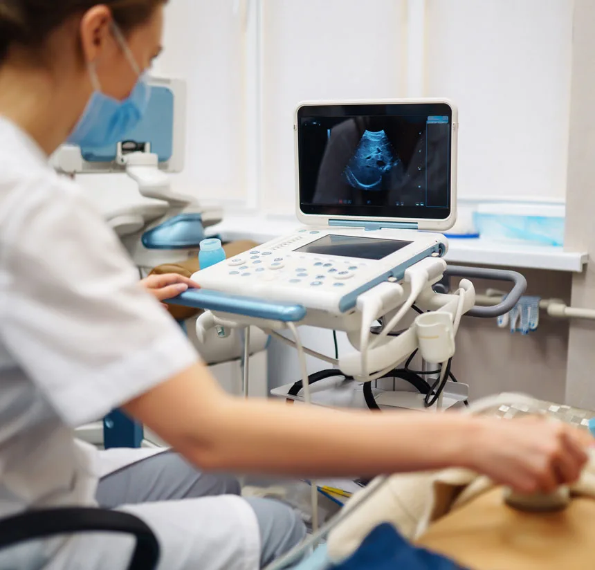 A healthcare professional performs an ultrasound scan on a patient, using a handheld device on the patient’s abdomen while viewing the results on a monitor.
