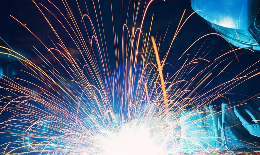 A person wearing protective gear and a helmet is welding metal, creating bright sparks and blue light against a dark background.