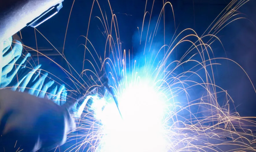A person wearing protective gear welds metal, creating bright blue light and orange sparks that fly outward in various directions against a dark background.