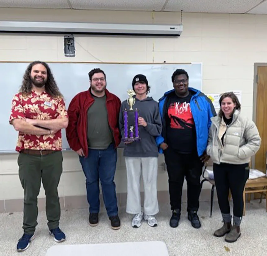 Five people stand indoors in front of a whiteboard, smiling at the camera. One person in the middle holds a large purple and gold trophy. They appear to be celebrating an achievement.