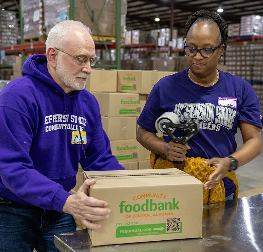 Two volunteers in purple shirts pack boxes labeled foodbank of Central Alabama in a warehouse. One person tapes a box while the other holds it steady. Stacks of boxes are visible in the background.