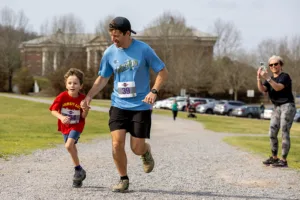 A man and young boy, both wearing race bibs, run together on a gravel path as a woman in the background smiles and takes a photo. Trees and parked cars are visible behind them.