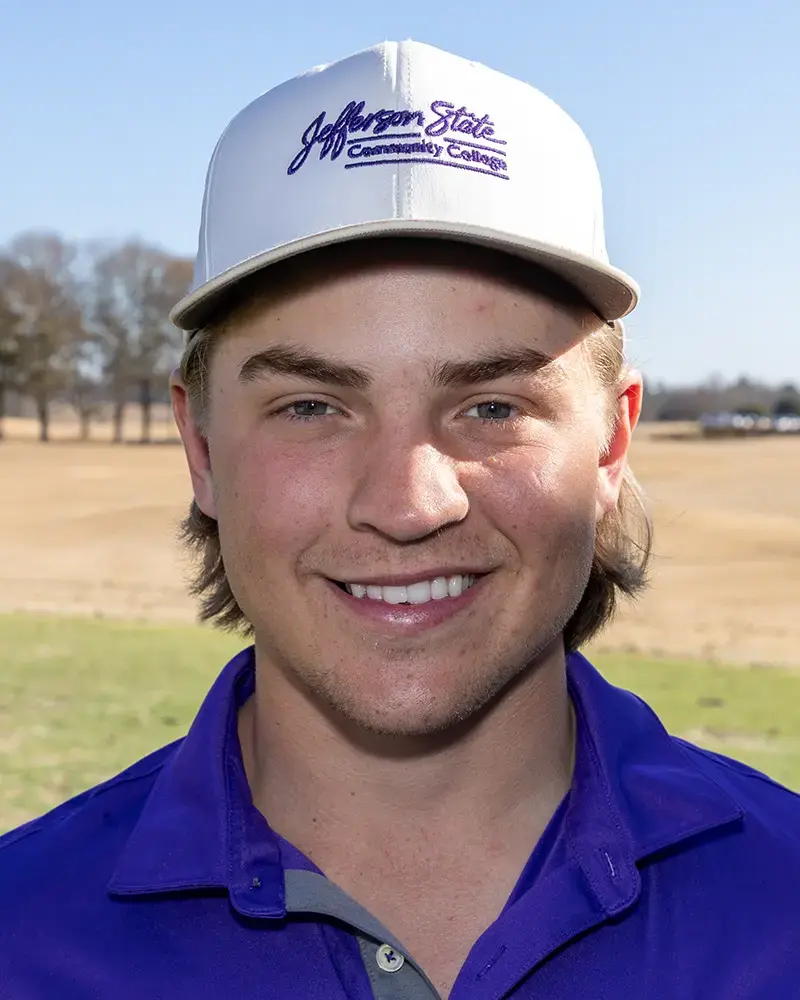 A young man wearing a white Jefferson State Community College cap and a blue polo shirt stands outdoors on a sunny day with a field and trees in the background, smiling at the camera.