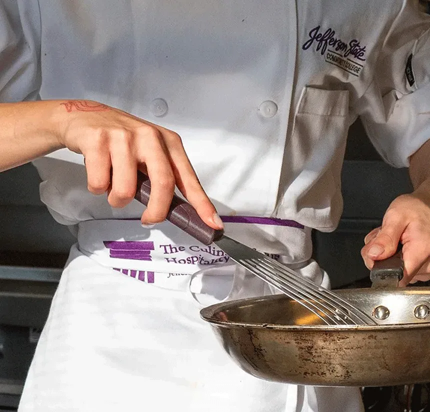 A person in a chef’s uniform stirs a pan with a metal whisk. The uniform reads “Jefferson State Culinary Institute” and sunlight illuminates their hands and the pan.