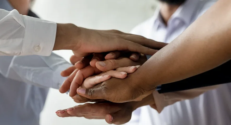 A group of people standing in a circle place their hands together, symbolizing teamwork.