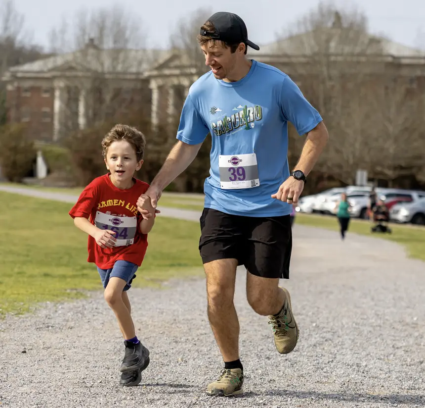 An adult and a child, both wearing race bibs, run together on a gravel path in a park-like setting. The child is smiling and wearing a red shirt, while the adult wears a blue shirt and a black cap.