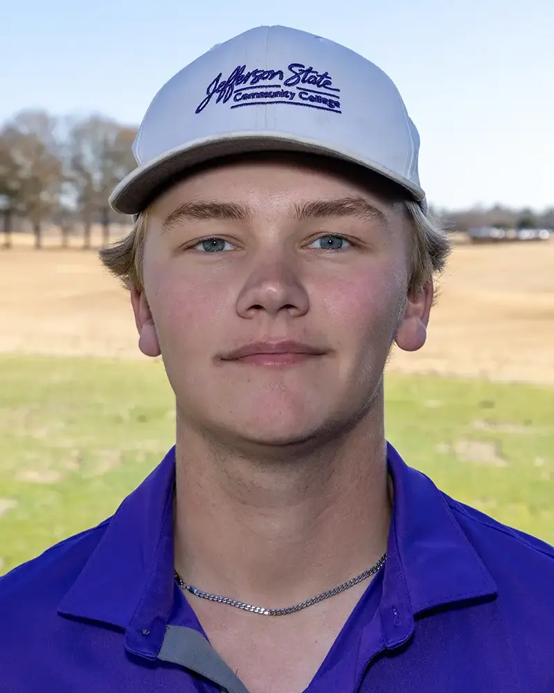 A young man wearing a white Jefferson State Community College cap and a purple polo shirt stands outdoors on a grassy field, looking at the camera with a neutral expression.