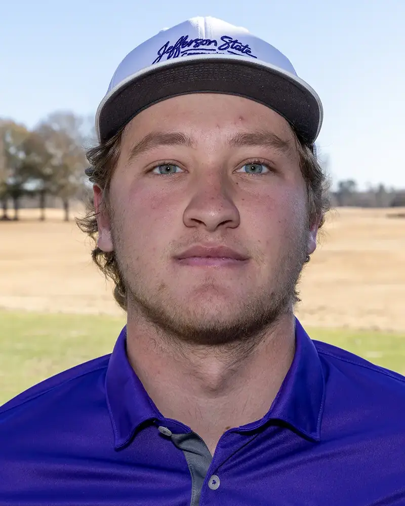 A young man wearing a white Jefferson State Community College cap and a blue collared shirt stands outdoors on a sunny day, with a grassy field and trees in the background.