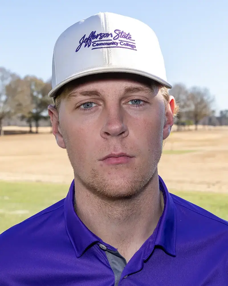 A young man wearing a purple Jefferson State Community College polo and a matching white cap stands outdoors on a golf course, looking directly at the camera with a neutral expression.