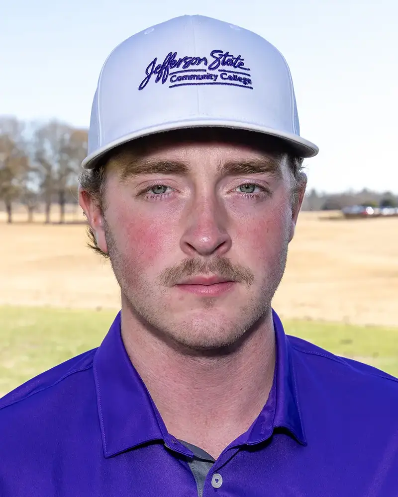 A young man in a white Jefferson State Community College cap and a purple collared shirt stands outdoors on a golf course, looking directly at the camera with a neutral expression.