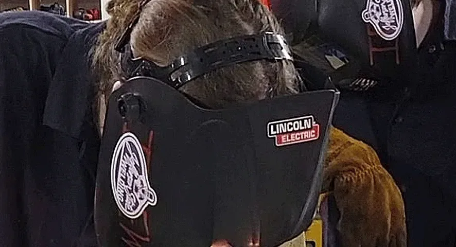 Two people wearing protective welding masks and gloves are working closely together, focused on welding a piece of metal in an industrial setting. Red safety curtains and equipment are visible in the background.
