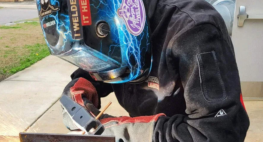 A person wearing a welding helmet and protective gear is welding metal outdoors, with bright sparks flying from the welding tool.