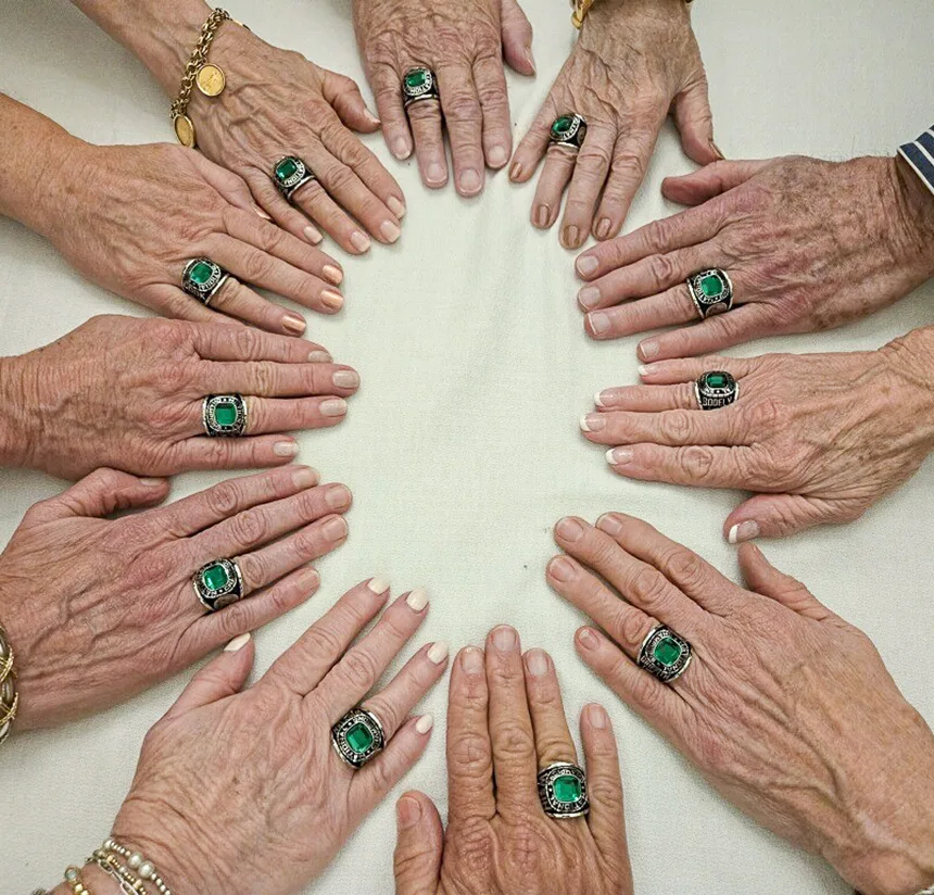 A circle of hands with visible signs of aging, each wearing a large green gemstone ring, is arranged on a cream-colored surface. Some wrists display gold bracelets and a wristwatch.