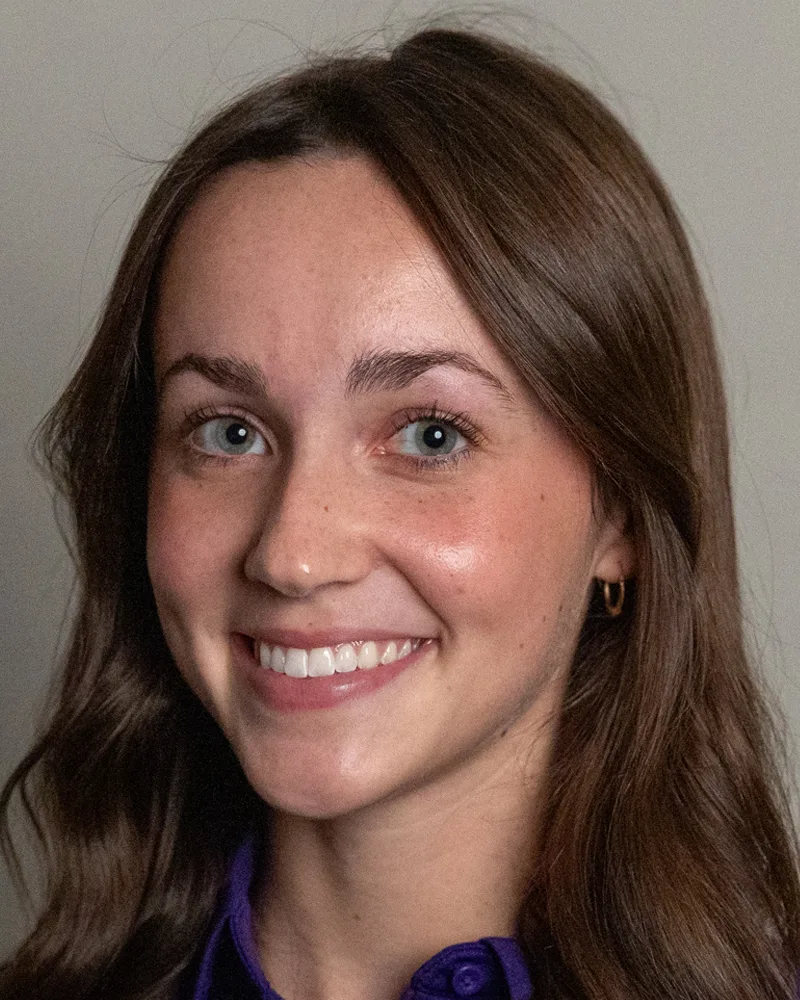 A young woman with long brown hair and blue eyes smiles at the camera. She is wearing a purple shirt and small hoop earrings, with a neutral background behind her.