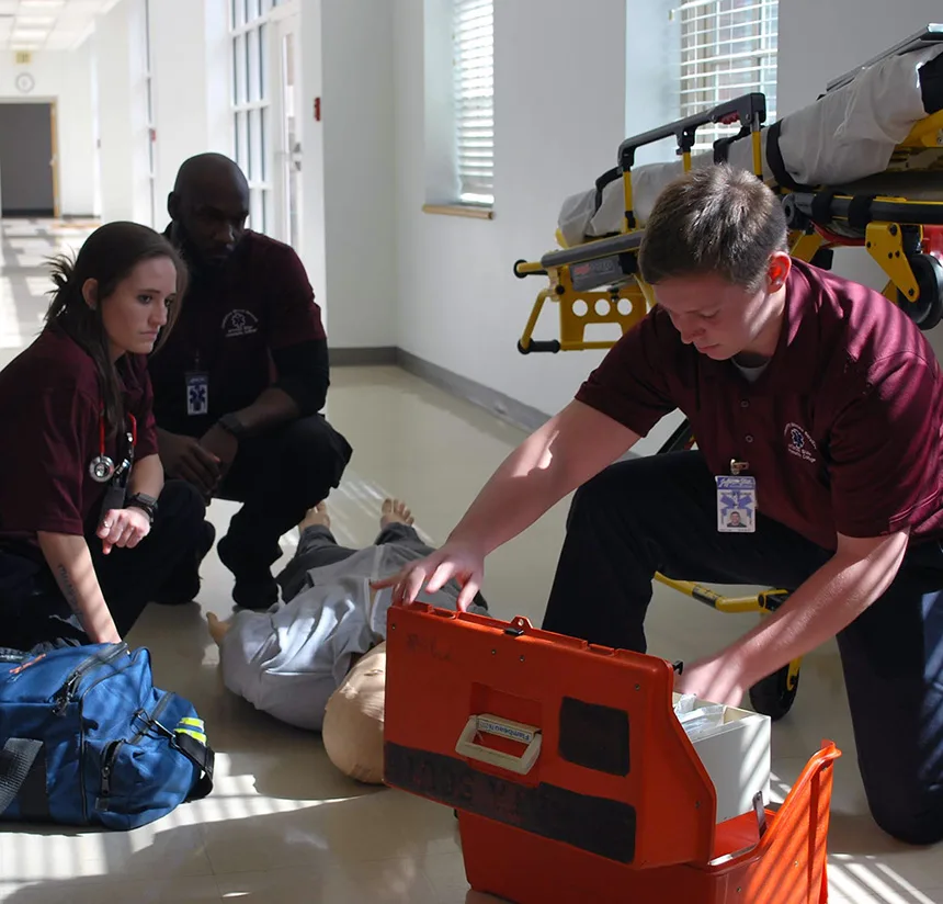 Three medical professionals attend to a mannequin lying on the floor in a hallway, with one opening an orange emergency kit and a stretcher in the background. Sunlight streams through the windows.