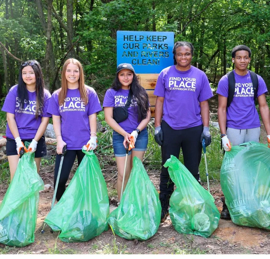 Five young people wearing purple Find Your Place shirts stand outdoors holding large green trash bags and litter pickers. Behind them is a sign that reads, Help Keep Our Parks and Rivers Clean.