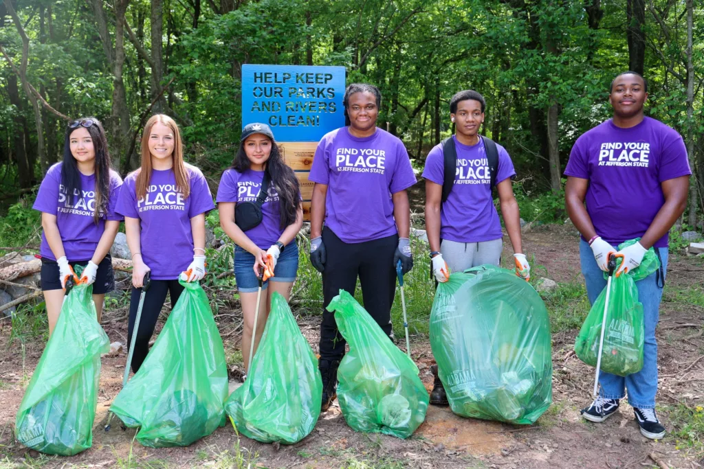Six young people in purple Find Your Place shirts stand outdoors, holding green trash bags and litter pickers, participating in a park clean-up. A sign behind them reads, Help Keep Our Parks and Rivers Clean!.