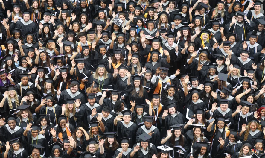 A large group of graduates in caps and gowns stand closely together, smiling and waving at the camera during a graduation ceremony.