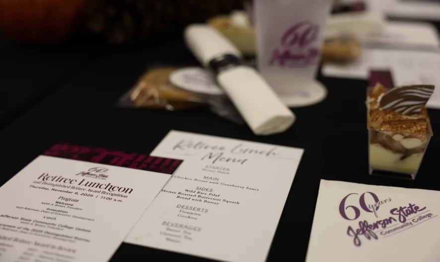 A close-up of a black table with printed menus, a rolled napkin, a cookie, a dessert cup, and a Jefferson State Community College cup, set for a retiree luncheon.