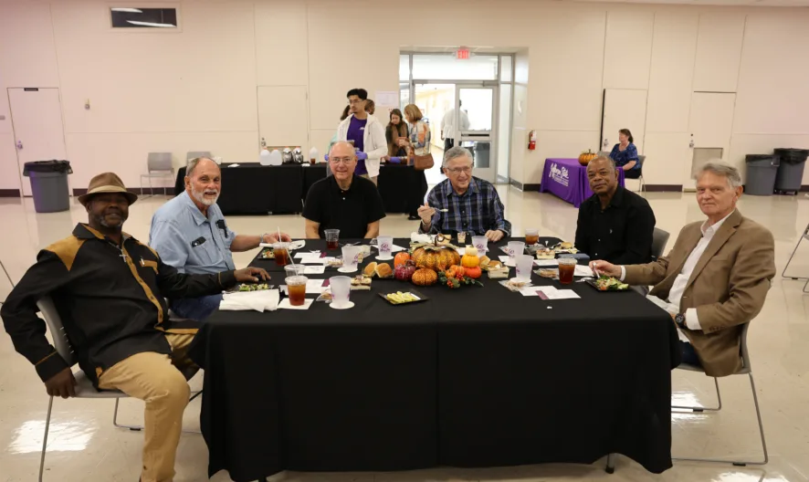 Six men sit around a rectangular table decorated with pumpkins and autumn-themed centerpieces, enjoying a meal together in a large, bright indoor space. Other people are visible in the background near a counter.