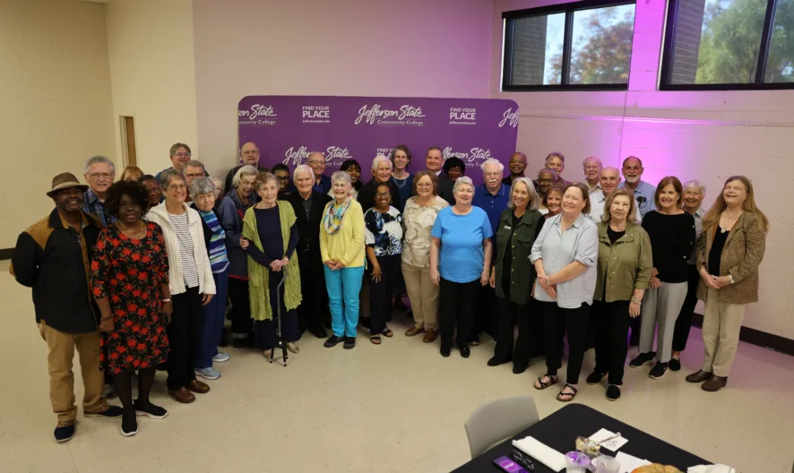 A large group of older adults stands together indoors, smiling at the camera. Behind them is a purple backdrop with “Jefferson State Community College” logos. The setting is a bright room with tables and windows.