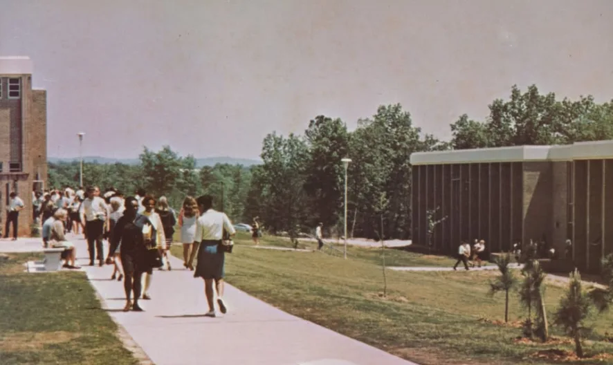 Students walk and gather along a wide sidewalk on a college campus, bordered by grassy areas and modern buildings, with trees and a blue sky in the background.