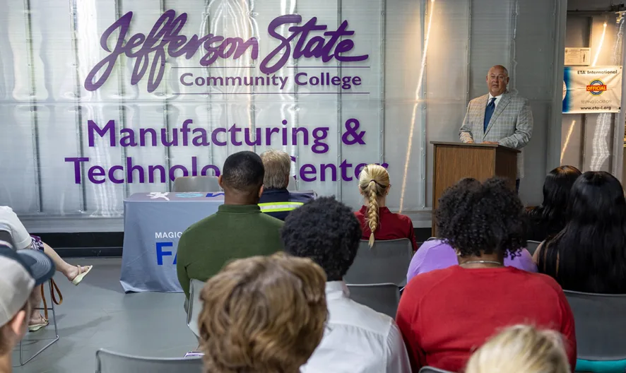 A group of people sits facing a speaker at a podium in front of a large sign reading Jefferson State Community College Manufacturing & Technology Center.