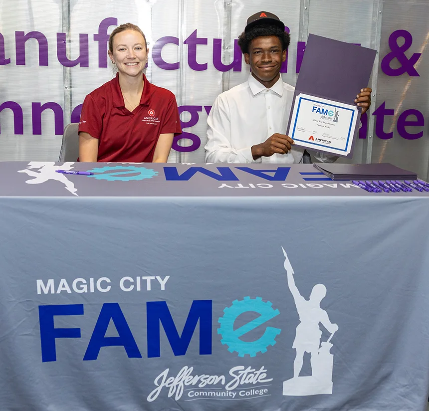 A smiling young man in a white shirt and cap holds a certificate next to a woman in a red shirt at a table with a Magic City FAME Jefferson State Community College banner. Both are seated in front of a branded backdrop.