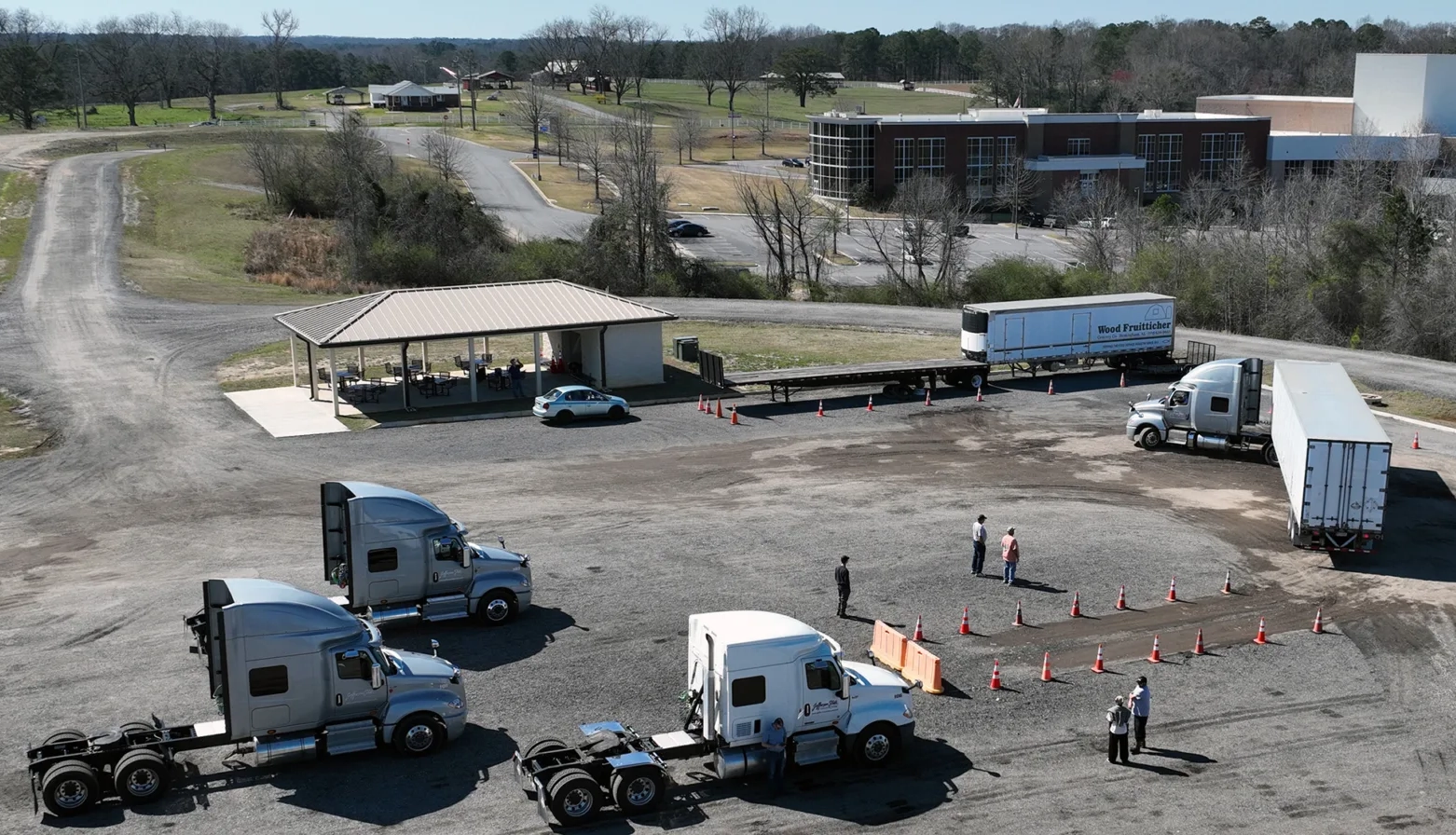 Aerial view of a truck driving training area with three semi-trucks, several people standing near traffic cones, a covered pavilion with tables, and a school building in the background.