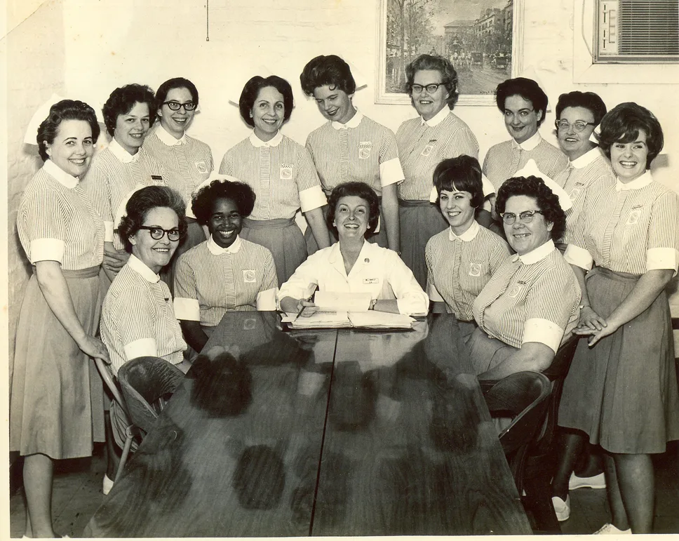 A group of thirteen women in matching uniforms stand and sit around a table, smiling, with one woman in white seated at the center. The background includes a painting, an air conditioner, and a brick wall.