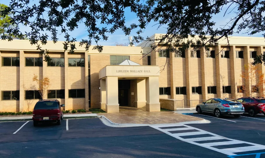 A beige brick building labeled Lurleen Wallace Hall stands behind a parking lot with several cars. Trees frame the image and the sky above is partly cloudy.